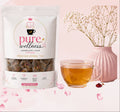 Pure Wellness herbal tea pouch shown on a soft pink background with decorative flowers, a pink vase, and a glass of amber tea. The package is white with pink and gold branding, and the text reads 'Cramps Don't Stand' and 'A Chance.